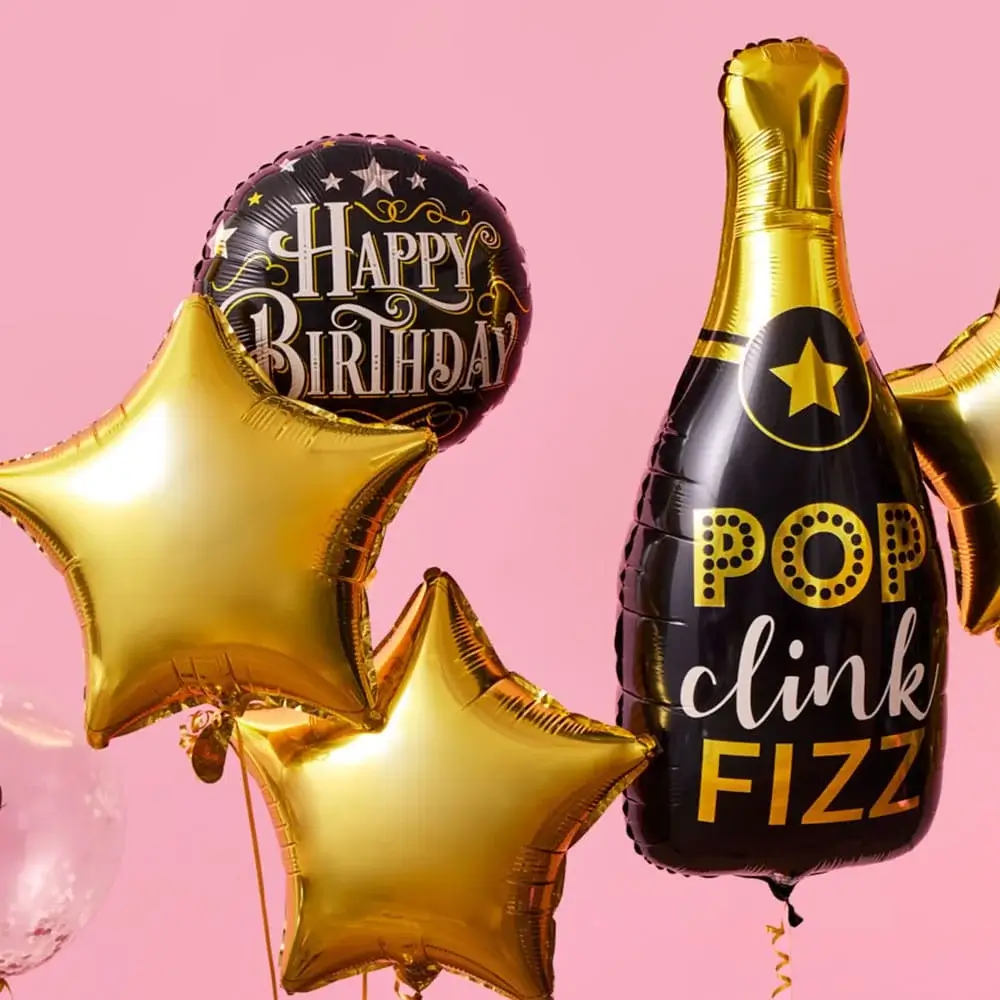 A collection of metallic balloons against a pink background, including two gold star-shaped balloons, a black round balloon with gold lettering that reads Happy Birthday, and a black champagne bottle-shaped balloon with gold accents and the words Pop Clink Fizz.